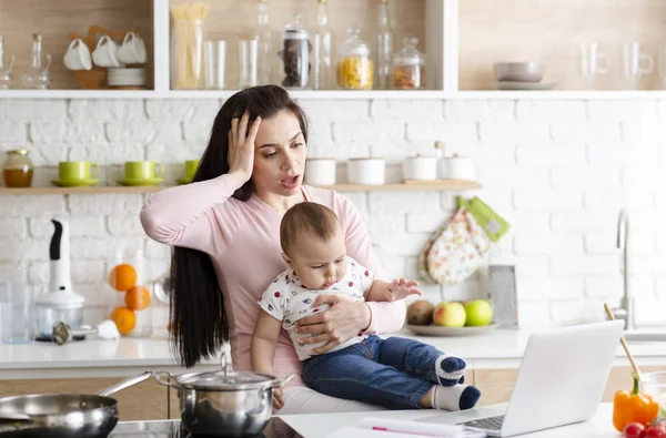 Desperate mother looking at laptop, working with baby in kitchen at home — Stock Photo, Image