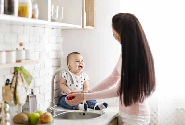 Entzückendes Baby sitzt auf dem Tisch in der Küche, während Mama Apfel wäscht — Stockfoto