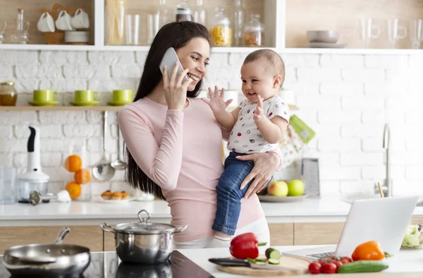 Mutter spricht mit Smartphone und hält entzückendes Baby in der Küche — Stockfoto