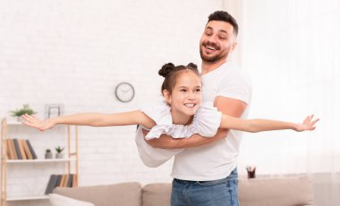 Baba, sevimli kızını uçak gibi yatarken tutuyor.