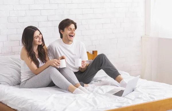Boldog pár élvezi a friss kávét, és nézi a laptopot az ágyban — Stock Fotó