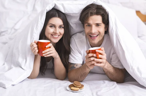 Веселая пара прячется под одеялом с чашками и печеньем — стоковое фото