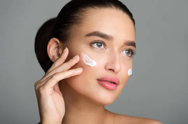 Hermosa mujer aplicando crema facial y mirando hacia otro lado —  Fotos de Stock