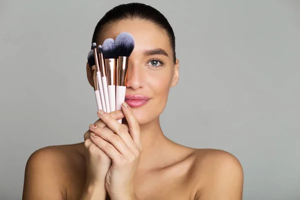 Vacker kvinna gömmer halva ansiktet bakom makeup borstar — Stockfoto