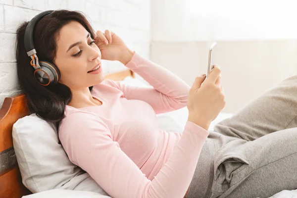 Mujer joven escuchando audiolibro con auriculares inalámbricos en la cama — Foto de Stock