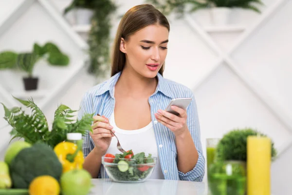 Giovane donna con telefono mangiare insalata fresca a tavola in cucina — Foto Stock