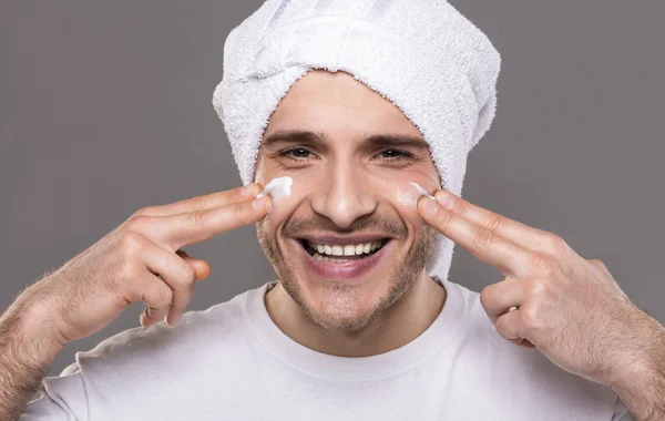 Duizendjarige kerel met handdoek op zijn hoofd, aanbrengen van crème op gezicht — Stockfoto