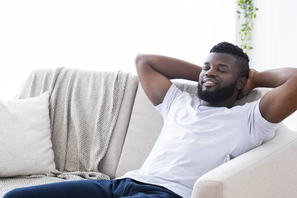 Cara africano feliz relaxando em casa, sentado no sofá com os olhos fechados — Fotografia de Stock