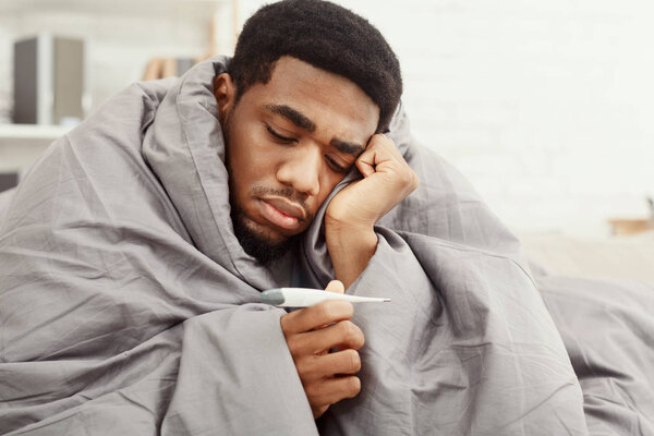 Sick african-american man wrapped in blanket holding thermometer