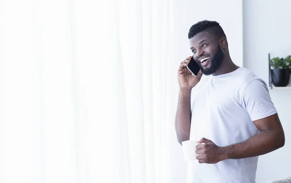 Fröhlicher schwarzer Mann, der mit dem Handy telefoniert, mit Kaffee zum Fenster schaut — Stockfoto