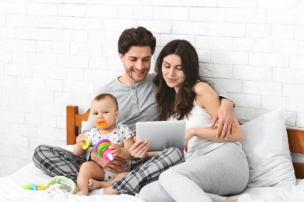 Pais navegando em tablet digital, sentados na cama com o bebê filho — Fotografia de Stock