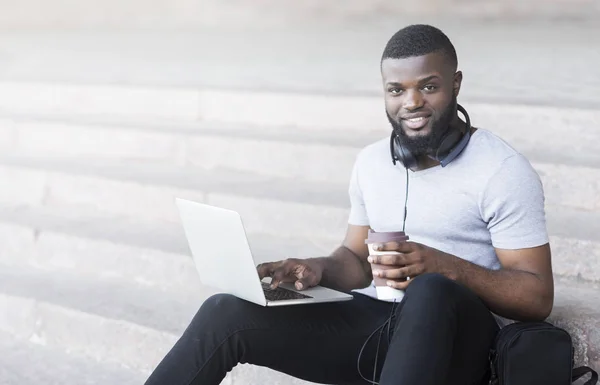 Smiling african student coding outdoor and drinking coffee