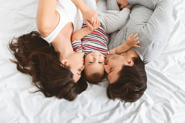Счастливые родители целуют милого маленького сына в постели — стоковое фото