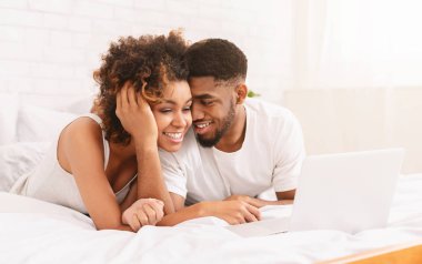 Genç siyah çift yatakta yatarken ve dizüstü bilgisayar kullanarak.