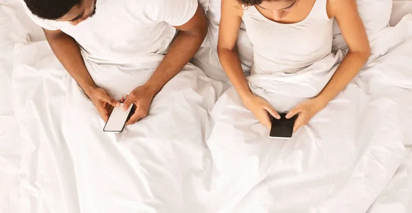 Millennial couple lying on bed back to back with smartphones