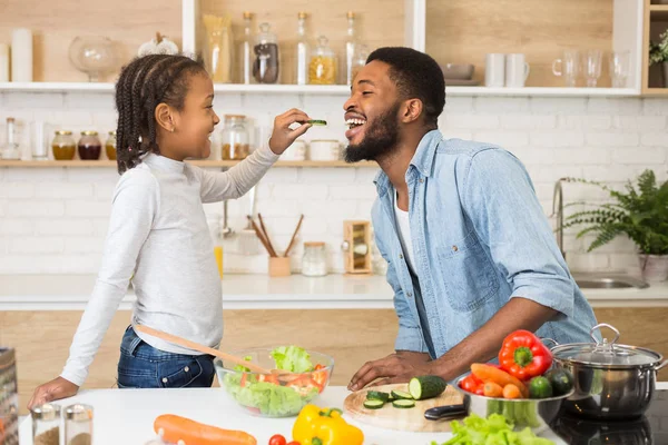 Afro girl está alimentando a su padre mientras cocina en la cocina —  Fotos de Stock
