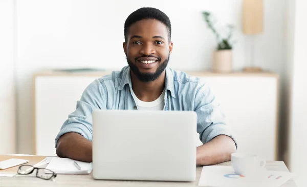 Africano usando el ordenador portátil, sonriendo y bebiendo café en la oficina —  Fotos de Stock