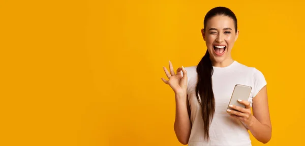 Vrouw houdt telefoon en Gesturing OK teken over gele achtergrond — Stockfoto