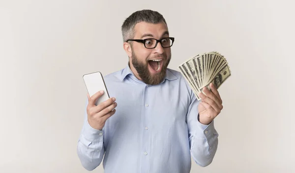 Vzrušený muž, který drží telefon a dívá se na hromadu peněz — Stock fotografie