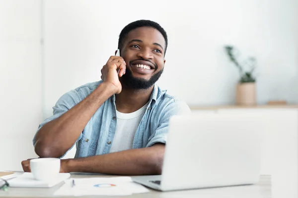 Hombre africano hablando por teléfono, trabajando en el ordenador portátil en la oficina — Foto de Stock