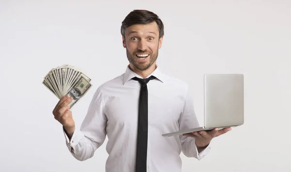 Beyaz arka plan üzerinde para ve dizüstü bilgisayar tutan heyecanlı genç adam — Stok fotoğraf