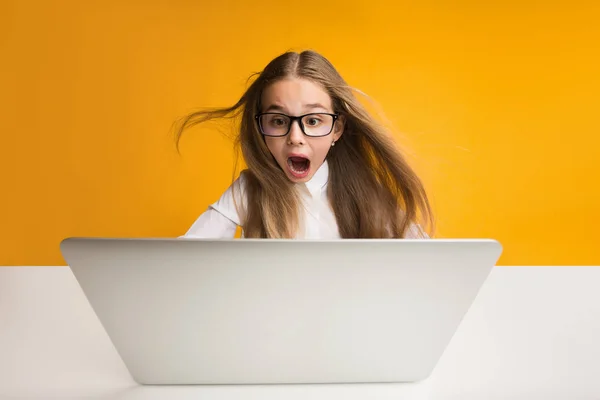 Sorprendido niña de la escuela haciendo la tarea en el ordenador portátil, fondo amarillo —  Fotos de Stock