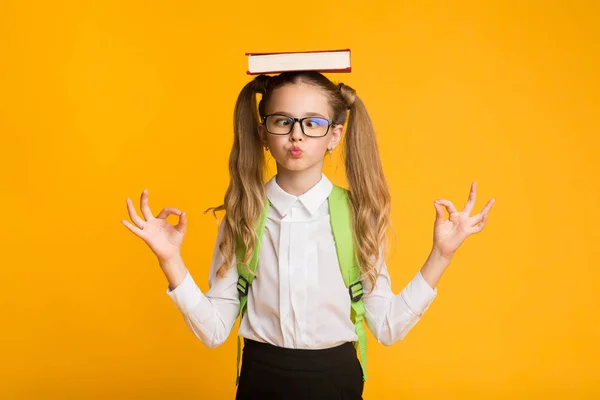 Basis school meisje mediteren houden boek op hoofd, gele achtergrond — Stockfoto