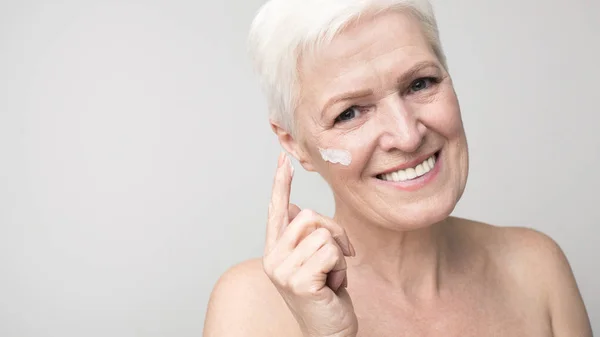 Donna anziana che applica la crema viso sulla guancia — Foto Stock