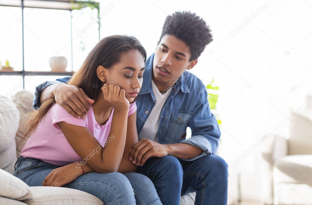 Young african guy comforting his sad girlfriend sitting on sofa