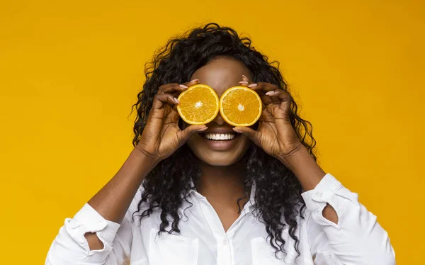 Chica negra alegre sosteniendo dos mitades de naranja cerca de los ojos — Foto de Stock