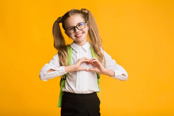 Weinig lachende schoolmeisje toont hart met handen op gele achtergrond — Stockfoto