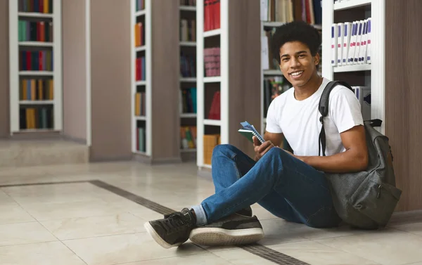 Estudante afro alegre sentado no chão da biblioteca ao lado de estantes — Fotografia de Stock