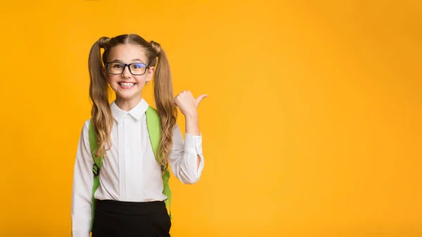 Alegre escola elementar menina apontando polegar no copyspace no amarelo — Fotografia de Stock