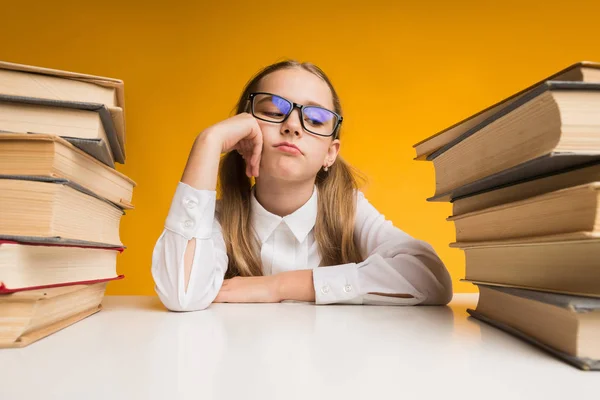 Chica aburrida de la escuela primaria sentada entre pilas de libros, fondo amarillo — Foto de Stock