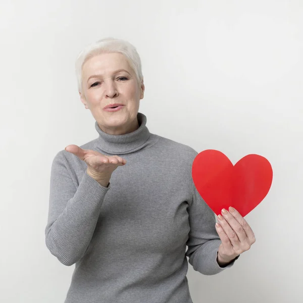 Όμορφη ηλικιωμένη γυναίκα που κρατά κόκκινη χάρτινη καρδιά και ανατινάζοντας φιλιά — Φωτογραφία Αρχείου