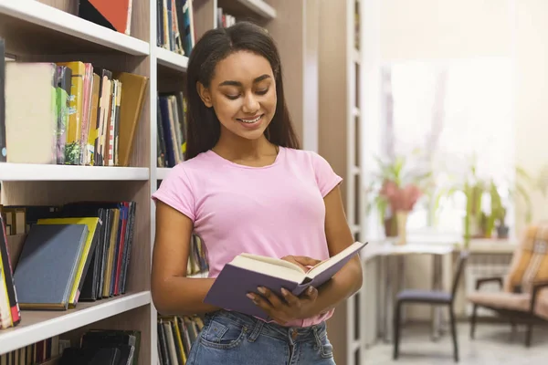 Sonriente niña afroamericana estudiante leyendo libro entre estanterías —  Fotos de Stock