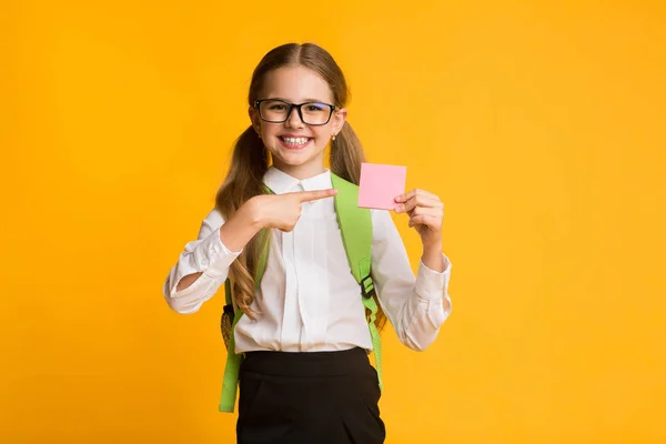Veselá školní dívka ukazující prstem na prázdnou kartu, žluté pozadí — Stock fotografie