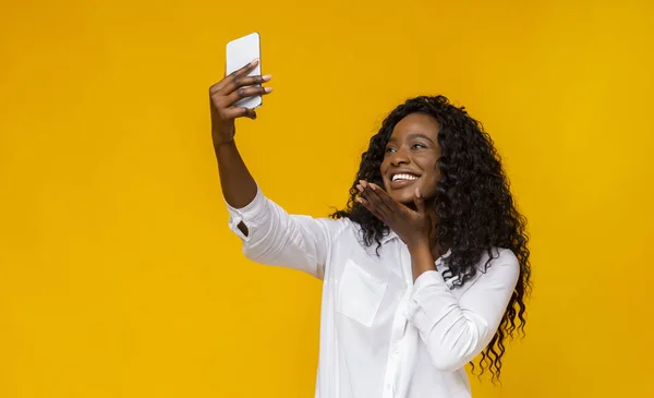 Бодрая черная девушка держит мобильник и делает селфи — стоковое фото