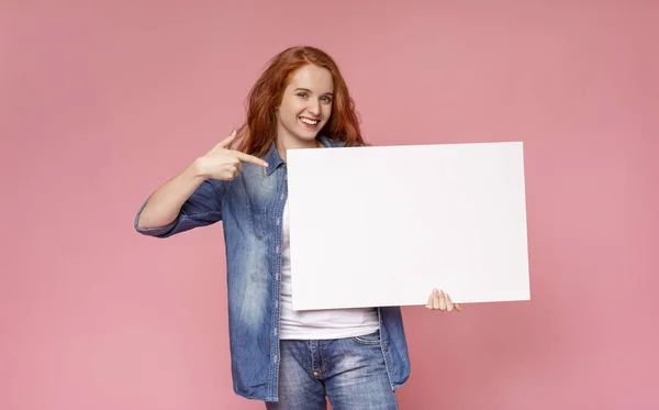 Menina de cabelos vermelhos segurando placa de publicidade em branco e apontando sobre ele — Fotografia de Stock