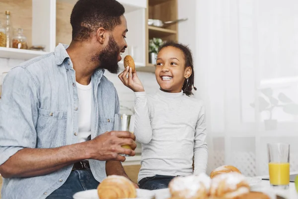 Alegre negro chica alimentación su papá, sentado juntos en cocina —  Fotos de Stock