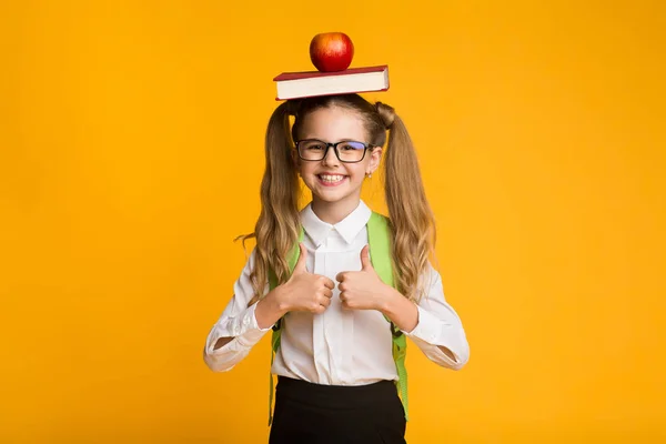 Studentessa gesturing pollice-su holding libro e mela su testa — Foto Stock
