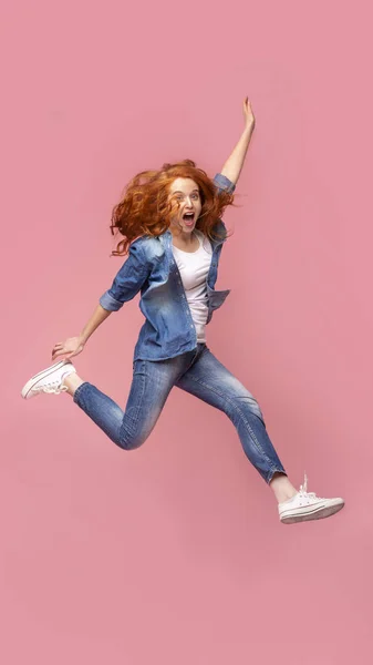 Позитивная рыжая девушка прыгает через розовый фон студии — стоковое фото