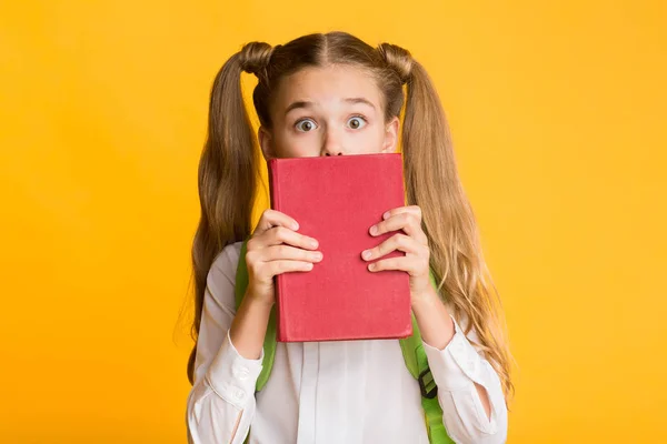 Vystrašená školní dívka skrývající tvář za knihou, žluté pozadí — Stock fotografie