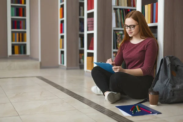 創造的な女子学生がエッセイを書き、図書館の床に座って — ストック写真