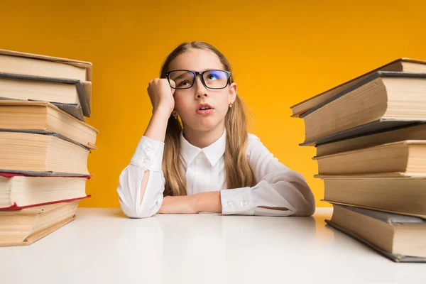 Colegiala descontenta sentada en la mesa entre pilas de libros, fondo amarillo — Foto de Stock