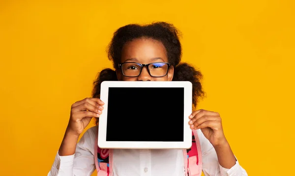 Colegiala afroamericana escondiéndose detrás de la pantalla en blanco de la tableta, fondo amarillo — Foto de Stock