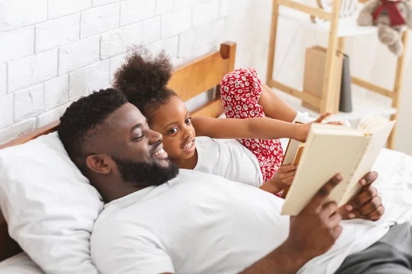 Zorgzame Afrikaanse papa lezen boek aan zijn dochter in de slaapkamer — Stockfoto