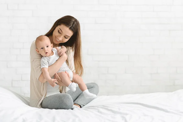 Mutlu anne meraklı yeni doğan bebek tutarak, birlikte yatakta oturan — Stok fotoğraf