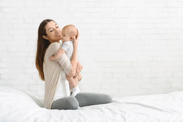 Lycklig tusenåriga kvinna sitter på sängen håller hennes bebis son — Stockfoto