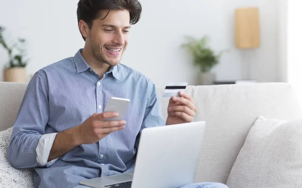Hombre sosteniendo la tarjeta de crédito y el teléfono celular sentado con el ordenador portátil en el interior —  Fotos de Stock